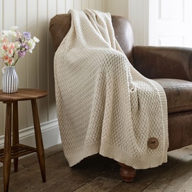 The Lyndon Company Cadenza Throw in cream, chunky knit texture with branded patch detail, draped over a leather armchair