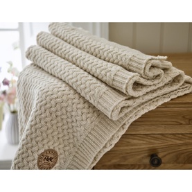 The Lyndon Company Cadenza Throw in cream, folded on a wooden surface, featuring chunky cable-knit texture and branded button detail.
