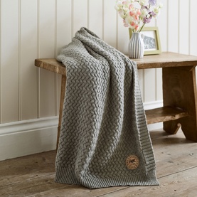 The Lyndon Company Cadenza Throw in grey, draped over a wooden bench, featuring a textured chevron knit pattern with striped edging.