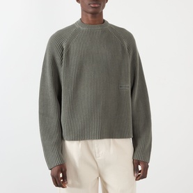 Model wearing About:Blank Logo Rib Knit Sweater in sage, crew neck, ribbed cotton with logo badge, styled with cream trousers