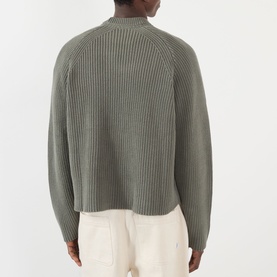 Model wearing About:Blank Logo Rib Knit Sweater in sage, back view showing ribbed cotton structure and raglan sleeves, styled with cream trousers.