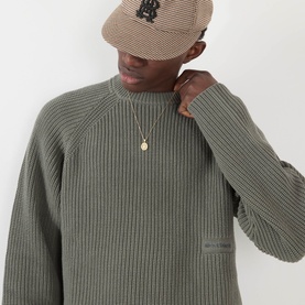 Model wearing About:Blank Logo Rib Knit Sweater in sage, ribbed cotton crew neck, styled with a beige cap and gold pendant necklace.