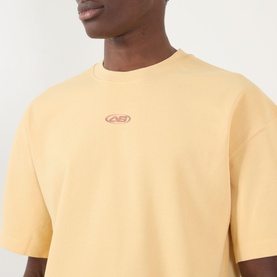 Model wearing About:Blank Raw Hem Ellipse T-Shirt in yellow, with small pink logo graphic at chest and relaxed fit.