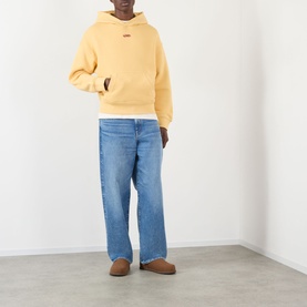 Model wearing About:Blank Arc Logo Hoodie in yellow with blue wide-leg jeans and tan suede slippers.