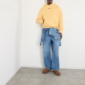 Model wearing About:Blank Arc Logo Hoodie in yellow with blue wide-leg jeans and a denim shirt tied at the waist, brown suede boots.