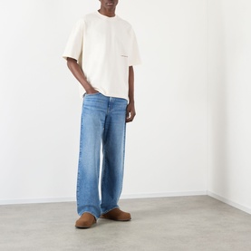 Model wearing About:Blank Oblique Logo T-Shirt in oat, oversized fit with round neck, styled with wide-leg blue jeans and tan suede shoes.
