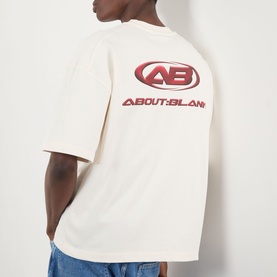 Model wearing About:Blank Oblique Logo T-Shirt in oat, rear view showing red oval AB logo print and brand name graphic on back.