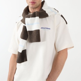Model wearing About:Blank oat cotton T-shirt with blue logo, styled with a brown and white striped knit scarf.