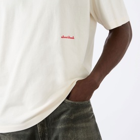 Model wearing About:Blank Haus Logo T-Shirt in oat with red logo print, styled with washed grey denim jeans.