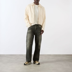 Model wearing About:Blank Haus Logo T-Shirt in oat/red, styled with a cream zip jacket and dark khaki wide-leg jeans and beige trainers.