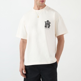 Model wearing About:Blank oat cotton T-shirt with black monogram chest graphic, styled with black trousers and gold necklace.