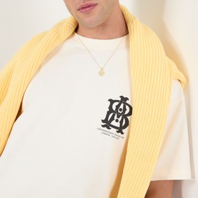Model wearing About:Blank oat cotton T-shirt with monogram chest graphic, styled with a yellow ribbed knit draped over shoulders.