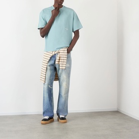 Model wearing About:Blank Box Logo T-Shirt in tourmaline/ecru, styled with distressed jeans and a striped shirt tied at the waist.