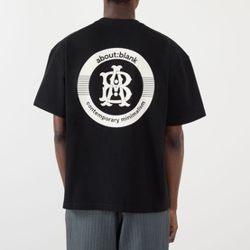 Model wearing About:Blank black cotton T-shirt, back view showing circular logo graphic print, styled with grey trousers.