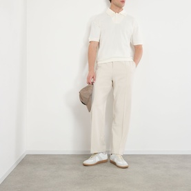 Model wearing white knit short-sleeve polo and cream wide-leg trousers, carrying a grey cap, styled with white leather trainers.