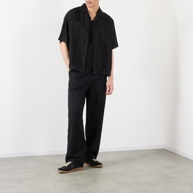 Model wearing black short-sleeve open-collar shirt with tonal pattern, styled with matching black wide-leg trousers and black trainers