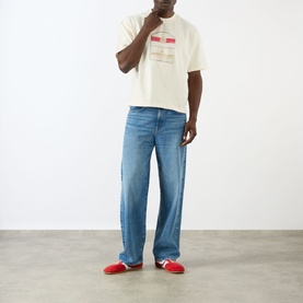 Model wearing About:Blank Graphic Logo T-Shirt in oat, crew neck, dropped shoulders, styled with blue wide-leg jeans and red trainers.