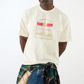 Model wearing About:Blank Graphic Logo T-Shirt in oat with red and gold front print, styled with plaid shirt tied at waist and blue jeans.