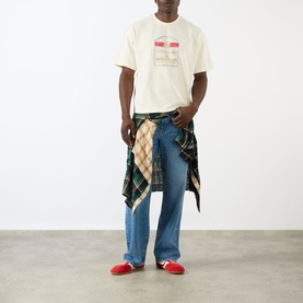 Model wearing About:Blank Graphic Logo T-Shirt in oat, styled with blue jeans and a plaid shirt tied at the waist, red trainers.