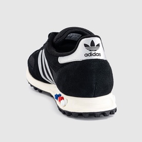 Adidas LA Trainer shoe in black suede with white three-stripe detail, cream midsole, and multicolour heel tab, rear angle view.