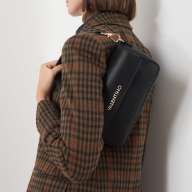 Model carrying Mario Valentino Alexia Flap Crossbody Bag in black with gold-tone logo lettering, worn as shoulder bag over brown checked coat