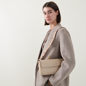 Model carrying Mario Valentino Alexia Flap Crossbody Bag in cream, with gold-tone hardware and logo lettering, worn crossbody over a beige coat.