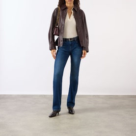 Model wearing dark brown leather zip-up jacket, styled with cream top, dark blue straight-leg jeans, and black heeled mules, carrying a burgundy bag