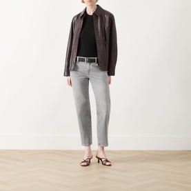 Model wearing dark brown leather jacket with black top, grey straight-leg jeans, brown belt, and strappy heeled mules