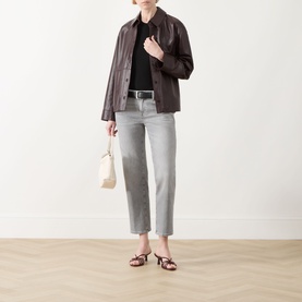 Model wearing dark brown leather jacket with black top, grey straight-leg jeans, and red strappy sandals, carrying a cream tote bag