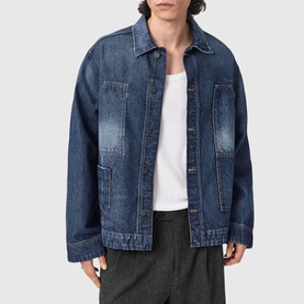 Model wearing All Saints dark indigo oversized denim jacket, open, with white T-shirt and grey trousers visible beneath.