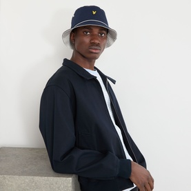 Model with Lyle & Scott Piped Logo Bucket Hat in Dark Navy, wide brim with tonal piping and gold eagle logo, styled with navy zip-up jacket