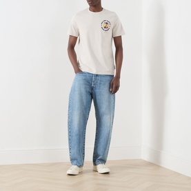 Model wearing Lyle & Scott Logo Nature Graphic T-Shirt in Putty with small chest graphic, styled with light blue wide-leg jeans and white trainers.