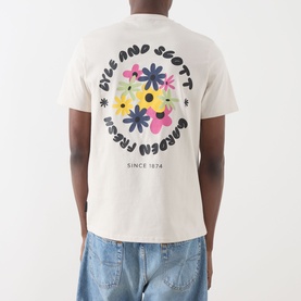 Model wearing Lyle & Scott Logo Nature Graphic T-Shirt in Putty with floral garden graphic on back, paired with light blue jeans.
