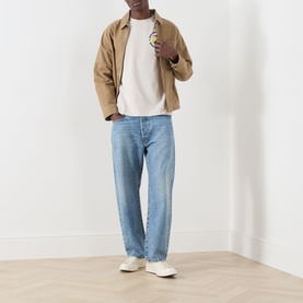 Model wearing Lyle & Scott Logo Nature Graphic T-Shirt in Putty with tan jacket, light blue jeans, and white trainers.