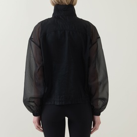 Model wearing Spanx jacket in Very Black, shown from behind, featuring sheer organza sleeves and high neck, paired with black trousers