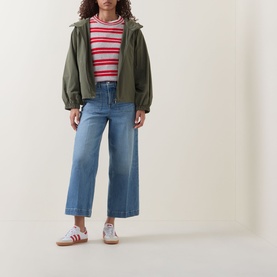 Model wearing olive green zip-up hooded jacket, red striped top, wide-leg blue jeans and white trainers with red stripes