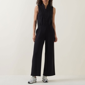 Model wearing Spanx AirEssentials&reg; Half-Zip Jumpsuit in Very Black, sleeveless with shawl collar and wide-leg fit, styled with white trainers.