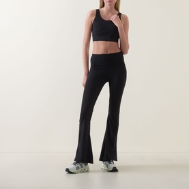 Model wearing Spanx Sheer Panel Sports Bra in Very Black with matching black flared leggings and grey trainers.