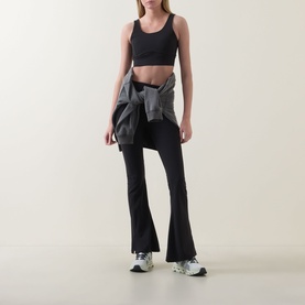 Model wearing Spanx Sheer Panel Sports Bra in Very Black with black flare trousers and a grey jacket tied at the waist.