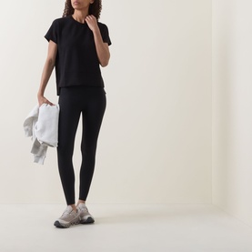 Model wearing Spanx AirEssentials T-Shirt in Very Black with matching black leggings and grey trainers, holding white jacket.