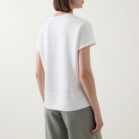 Model wearing Spanx AirEssentials&reg; T-Shirt in Powder, back view showing dipped hemline and short sleeves, styled with grey joggers.