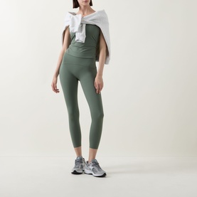 Model wearing Spanx BoostUp Shelf Tank in Eucalyptus, styled with matching green leggings, white draped top, and grey trainers.