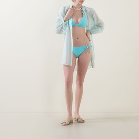 Model wearing Naia Beach Ursula Aqua Side-Tie Bikini Briefs with matching triangle top and sheer pale blue cover-up shirt and gold sandals.
