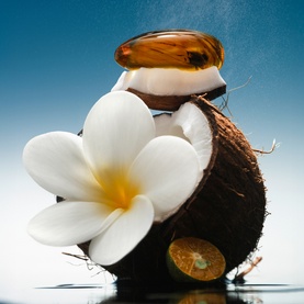 Phlur Island Swim Hair and Body Mist styled with a halved coconut, amber capsule, and white frangipani flower on a blue gradient background