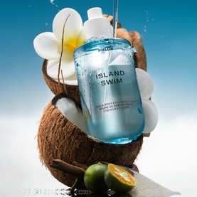 Phlur Island Swim Hair and Body Mist in blue bottle displayed in a halved coconut with tropical flowers and citrus fruit
