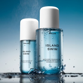 Phlur Island Swim Hair and Body Mist in two sizes, clear blue bottles with white caps, surrounded by water droplets on reflective surface.