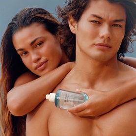 Model with Phlur Island Swim Hair and Body Mist; male model holds bottle while female model rests on his shoulder, both with sun-kissed skin