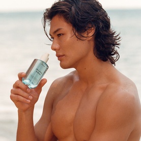 Model with Phlur Island Swim Hair and Body Mist, holding light blue spray bottle, shot at beach setting