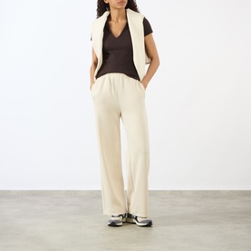 Model wearing Adanola Coffee Bean top styled with cream wide-leg trousers, white gilet, and grey trainers.