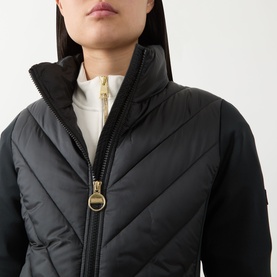 Model wearing Barbour International black quilted jacket with gold-tone zip and ring pull detail, styled over white top, high collar.